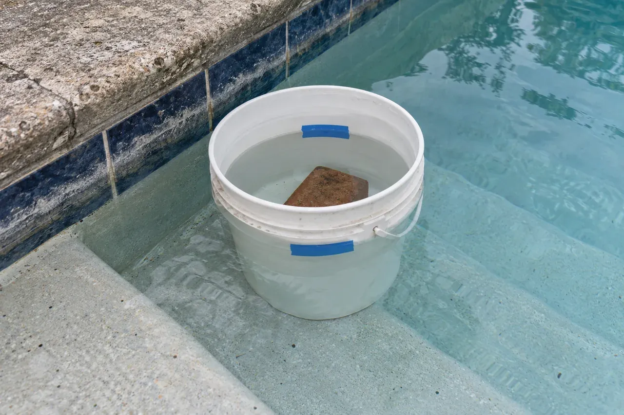 An unbranded white 5-gallon plastic bucket weighted with a red clay brick on a submerged plaster pool step